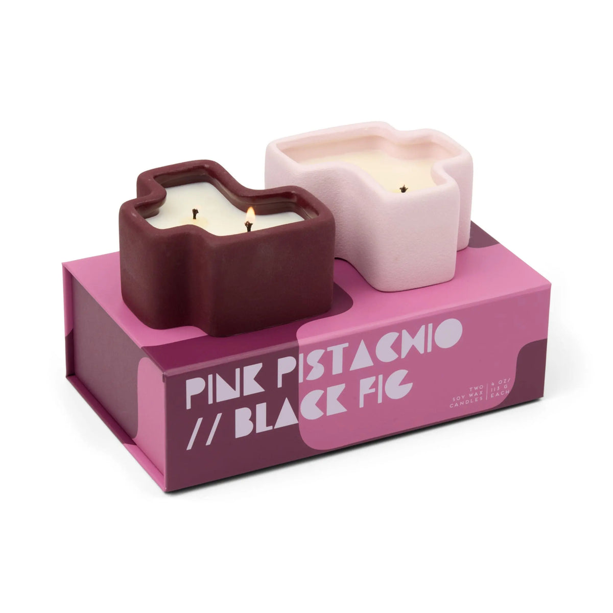 Two uniquely shaped candles—one in dark purple and one in light pink—are displayed on a pink box boldly labeled "PINE PISTACHIO // BLACK FIG," creating the eye-catching Pink Pistachio & Black Fig - Jigsaw 4 oz Candle Boxed Set.