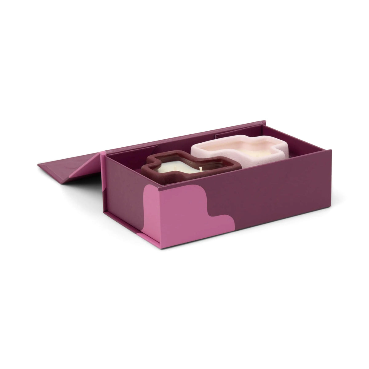 A purple box with pink accents and a wavy design on the side contains the Pink Pistachio & Black Fig - Jigsaw 4 oz Candle Boxed Set, featuring two abstract blob-shaped candles—one white and one purple.
