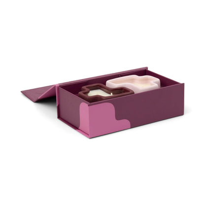 A purple box with pink accents and a wavy design on the side contains the Pink Pistachio & Black Fig - Jigsaw 4 oz Candle Boxed Set, featuring two abstract blob-shaped candles—one white and one purple.
