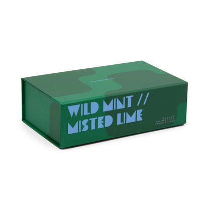 A green rectangular box featuring abstract shapes and bold blue text reading "WILD MINT // MISTED LIME" on the side, designed for the Wild Mint & Misted Lime - Jigsaw 4 oz Candle Boxed Set, suggesting a refreshing and thoughtfully curated ceramic candle pairing inside.
