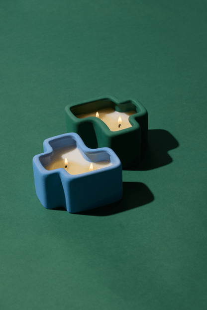 Two abstract-shaped candles—one in blue and one in green—are lit and positioned on a green background, each displaying three flames with shadows extending to the right. This eye-catching composition evokes the invigorating fragrances of the Wild Mint & Misted Lime - Jigsaw 4 oz Candle Boxed Set.