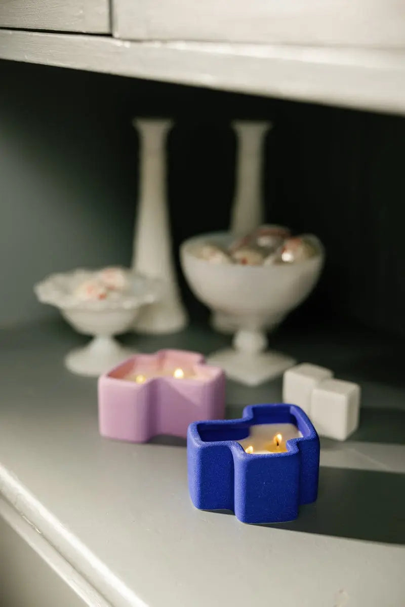 Two lit star-shaped candles from the Wisteria & Ember + Resin - Jigsaw 4 oz Candle Boxed Set, one in purple and one in blue, sit on a gray shelf. Their gentle floral fragrance fills the air, creating a serene ambiance. In the background, white ceramic candlesticks and bowls with decorative items complete the scene.