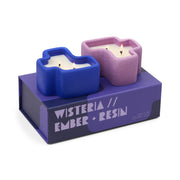 Two abstract-shaped candles—one blue and one purple—rest atop a purple box labeled "Wisteria & Ember + Resin - Jigsaw 4 oz Candle Boxed Set." Each candle features three wicks, their elegant forms side by side, inviting you to experience their delicate floral fragrance.