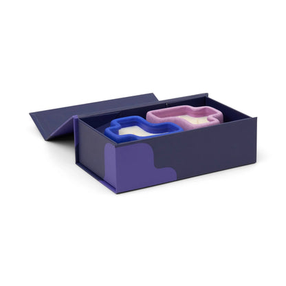 A navy blue box with purple accents, evocative of the Wisteria & Ember + Resin - Jigsaw 4 oz Candle Boxed Set, holds two shoe-shaped sponges—one in white and blue, the other in pink and purple—with its lid partially open.