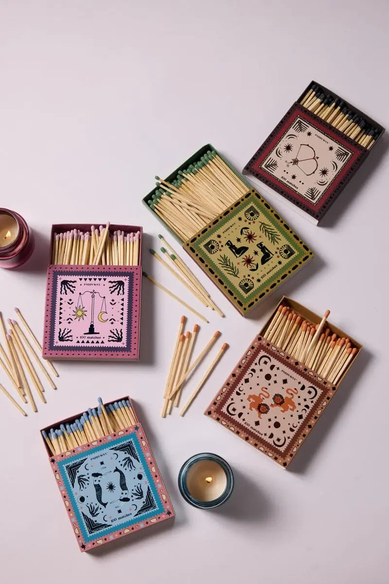 Bow & Arrow - Black Matches: Five decorative matchboxes, each with a unique design and colorful border, are arranged on a white surface beside lit candles and loose jumbo matches as the ideal candle accessory.