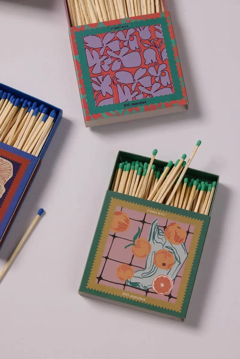 Three Oranges - Green Matches decorative matchboxes with colorful designs and green-tipped jumbo matches are displayed on a light surface as the perfect candle accessory. A single match is placed separately for added style.