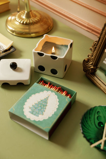 A Misted Lime - Lucky Dice 10 oz Candle in a white, die-shaped ceramic vessel with its lid beside it, next to an open box of matches featuring a Christmas tree illustration.