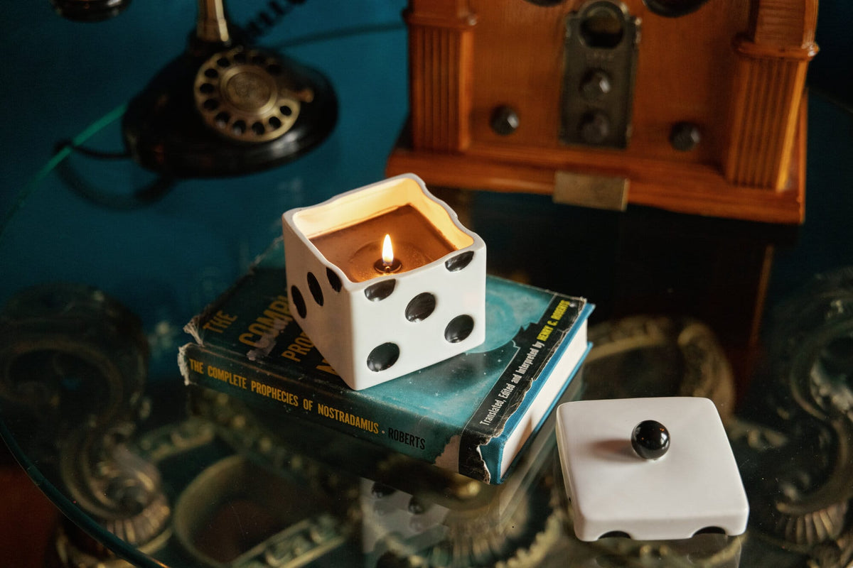 A Tobacco Patchouli - Lucky Dice 10 oz Candle in a white ceramic dice vessel sitting on top of an old book, with the matching lid resting nearby.