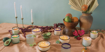 A table featuring the Salted Blue Agave - La Playa 9 oz Candle, fruit, flowers, avocados, a decorative vessel, a vase with dried palm leaves, and two tall candlesticks set against a light blue wall.
