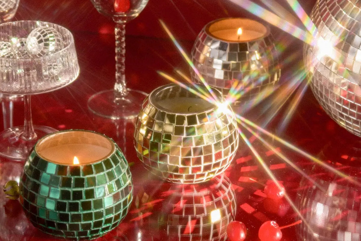 The Tobac & Pine - Magique Disco Ball 10 oz candle shines on a red reflective surface, with festive beads and glassware in the background, making this disco-inspired accent perfect for stylish home decor.