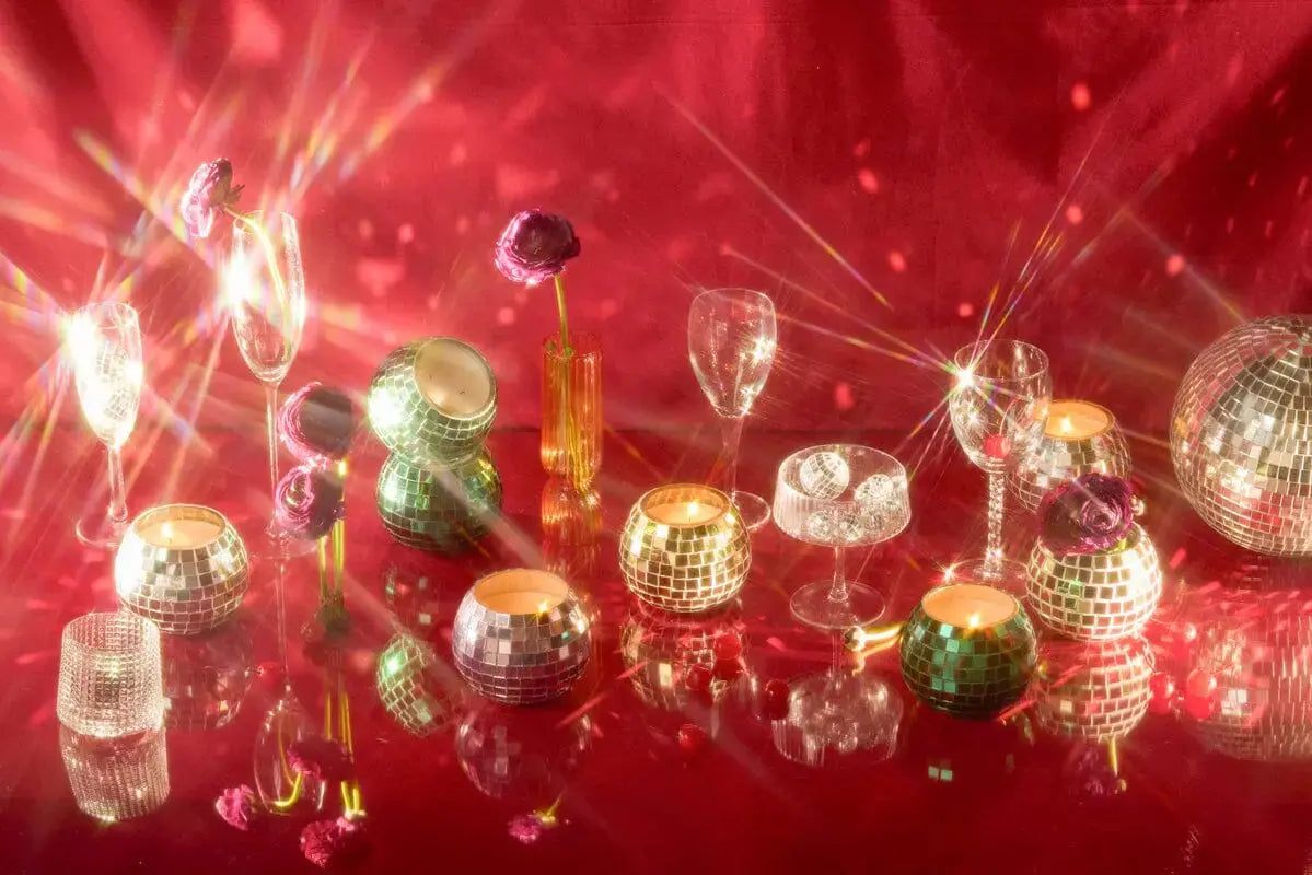 The Sparkling Bergamot - Magique Disco Ball 10 oz candle shines among glassware and flowers on a reflective red surface, with light rays and starbursts creating a festive and glamorous scene.