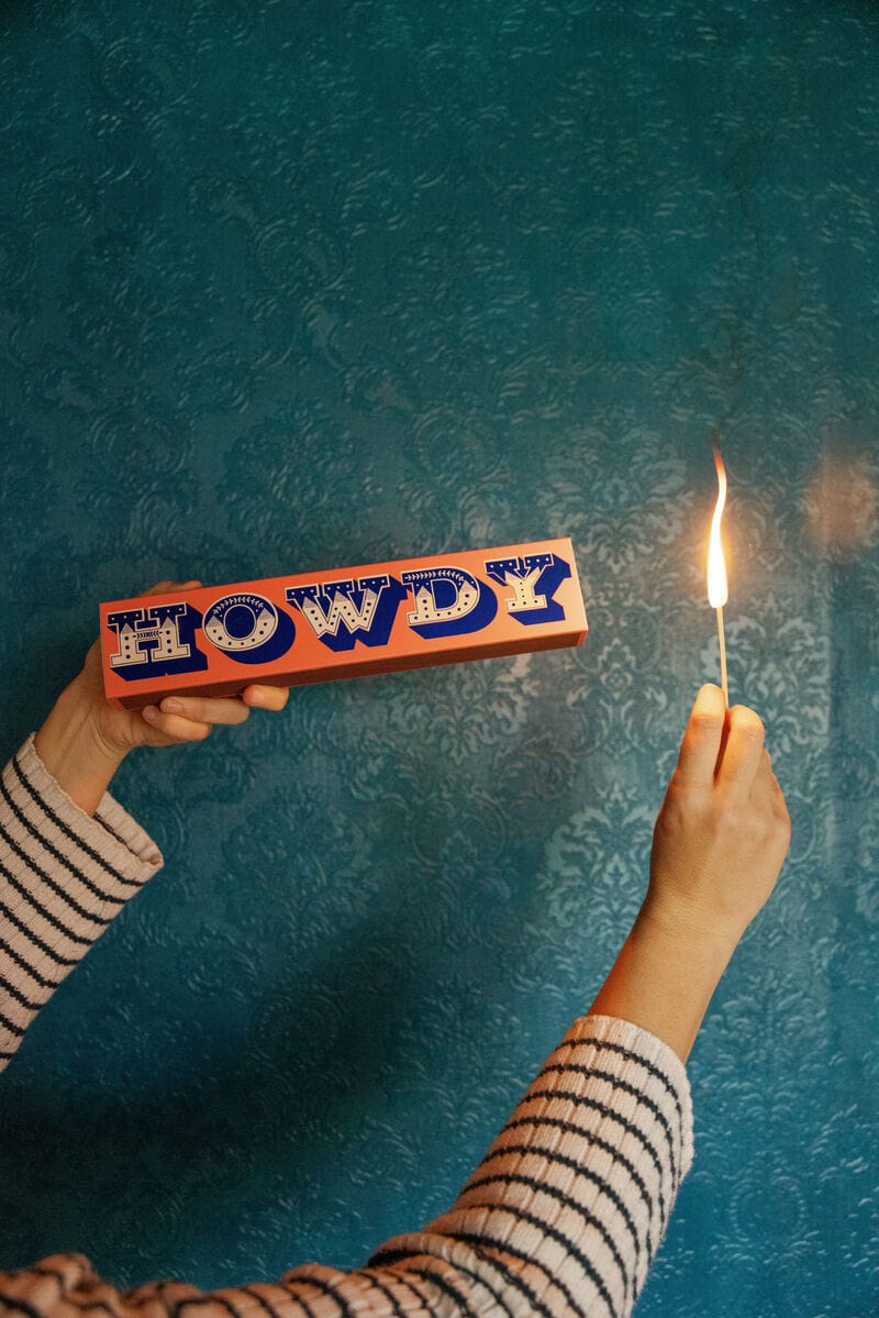 A person in a striped sweater holds a Howdy - Extra Long Boxed Matches in one hand and a lit match in the other against a textured teal wall.