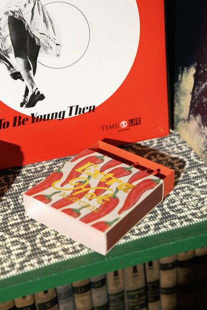 A red Extra Hot - Square Boxed Matches sits on a patterned surface, making a bold host gift in front of a red Time Life book with a black-and-white image of dancing legs.