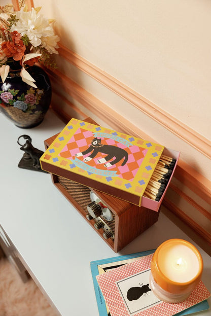 A colorful Bougie Cat - Large Rectangle Boxed Matches with a cat illustration sits on a radio beside a candle, books, figurine, and floral vase—perfect decor for cat lovers and match enthusiasts.