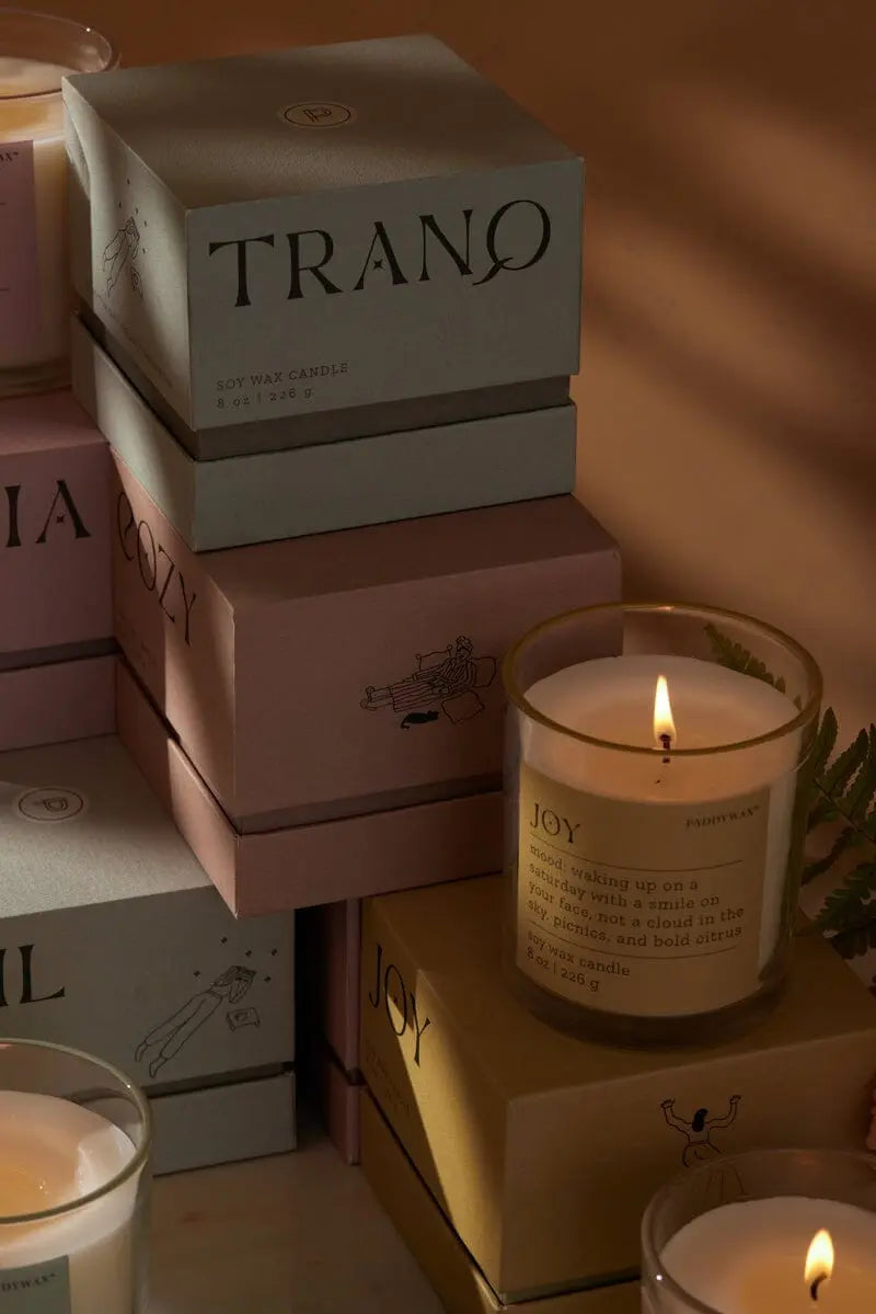 A lit Misted Lime "Joy" - Mood Collection 8 oz candle in a glass holder stands before stacked, muted candle boxes, glowing softly and warmly.