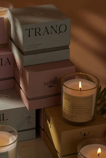 A lit Misted Lime "Joy" - Mood Collection 8 oz candle in a glass holder stands before stacked, muted candle boxes, glowing softly and warmly.