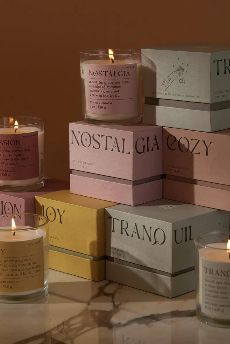 The Pepper + Plum "Nostalgia" Mood Collection 8 oz candle is displayed lit in a glass jar atop a colorful box, adding the perfect mood-setting touch to any space.