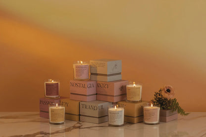The Misted Lime "Joy" 8 oz candle from the Mood Collection is displayed in a glass jar with other lit candles on pastel boxes with printed labels, set on marble against a warm-toned gradient and a small floral arrangement.