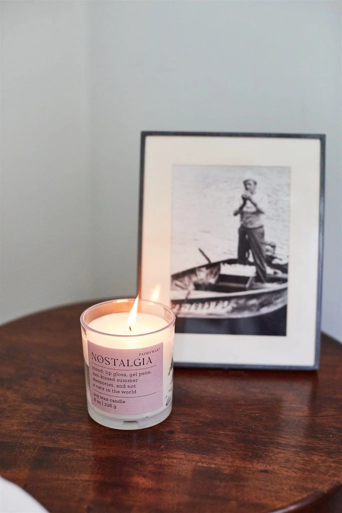 A Pepper + Plum "Nostalgia" Mood Collection 8 oz candle rests on a wooden table next to a framed black-and-white photo of someone in a rowboat.