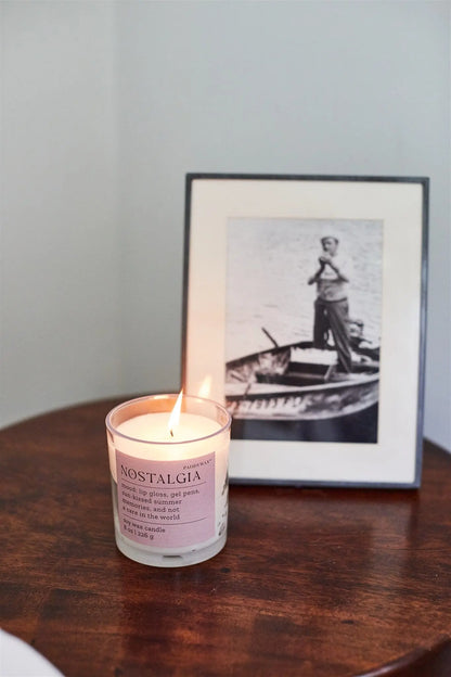 A Pepper + Plum "Nostalgia" Mood Collection 8 oz candle rests on a wooden table next to a framed black-and-white photo of someone in a rowboat.