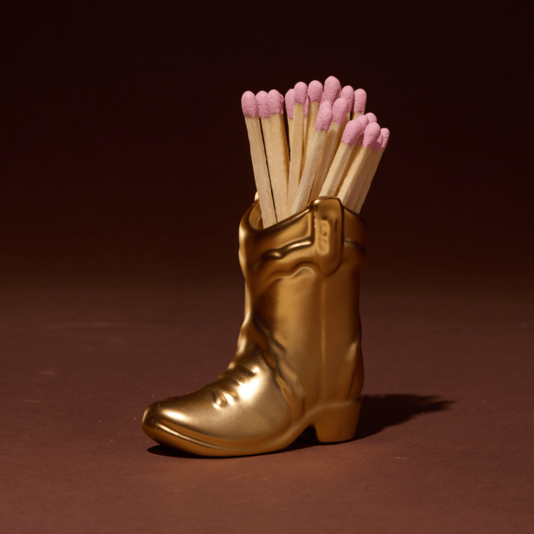 The Cowboy Boot Match Holder - Gold Metallic is a small ceramic cowboy boot finished in shimmering gold, holding a bunch of wooden matches with pink tips. Set against a dark brown background, it makes the perfect piece of Western home decor or a unique Nashville gift.