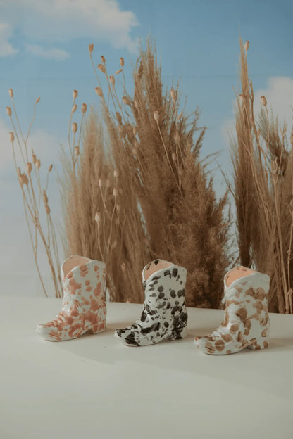 Three small ceramic boots in pink, black, and brown cow print sit on a white surface, capturing the charm of the Cactus Flower - Nashville Boot 6oz Candle, with tall dried grass and a bright blue sky in the background.