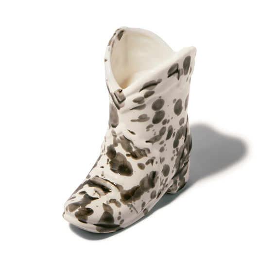 The Palo Santo Suede - Nashville Boot 6 oz Candle features a ceramic vessel shaped like a cowboy boot with black and brown patterns on white, casting a shadow to the right on a plain surface.