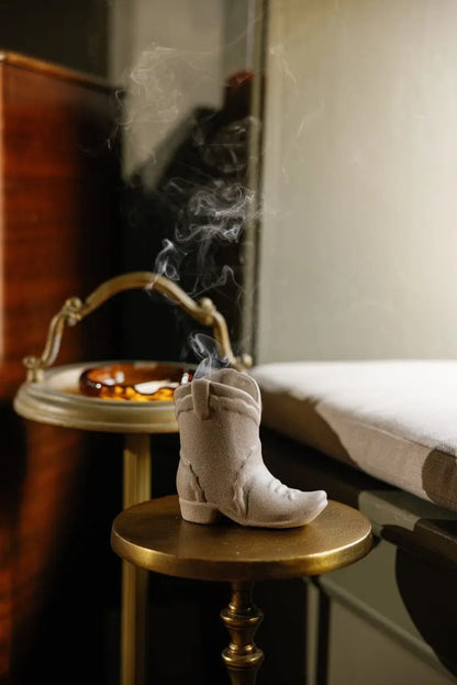 A Bergamot & Mahogany - Nashville Boot 6 oz Candle, shaped like a small cowboy boot, rests on a brass table with its gentle smoke curling upward—filling the air with warm, western-inspired fragrance. In the background, a tray displays a glass ashtray. The softly lit scene exudes cozy charm and pays homage to Nashville-inspired décor.