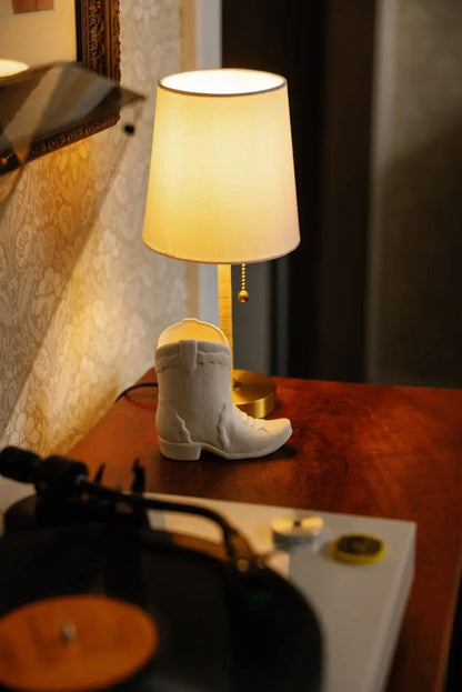 A lit Smoked Pistachio - Nashville Boot oz Candle and a small white cowboy boot decoration sit on a wooden surface near a record player, with a patterned wall in the background, creating a cozy, Nashville-inspired atmosphere.