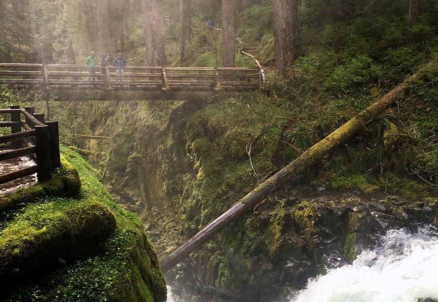 A wooden bridge spans a deep, lush canyon above a rushing river, surrounded by dense green forest and moss-covered rocks. Three people stand on the bridge, looking out over the scenery. Mist rises from the water below.