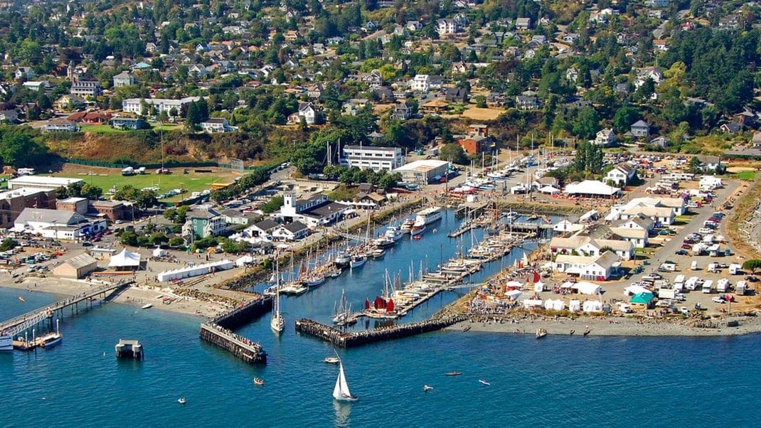Aerial view of a coastal marina with numerous sailboats docked, surrounded by white tents and buildings. A small sailboat is on the water, and a residential neighborhood is visible in the background.