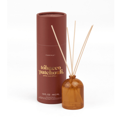 The Tobacco Patchouli - 1.5 oz Diffuser features a brown cylindrical container beside a petite amber glass bottle with wooden reeds, offering a charming and giftable fragrance option.