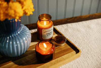 A lit Pumpkin & Spice candle and a small brown candle sit on a wooden tray beside a blue vase with yellow flowers, on a textured white surface.