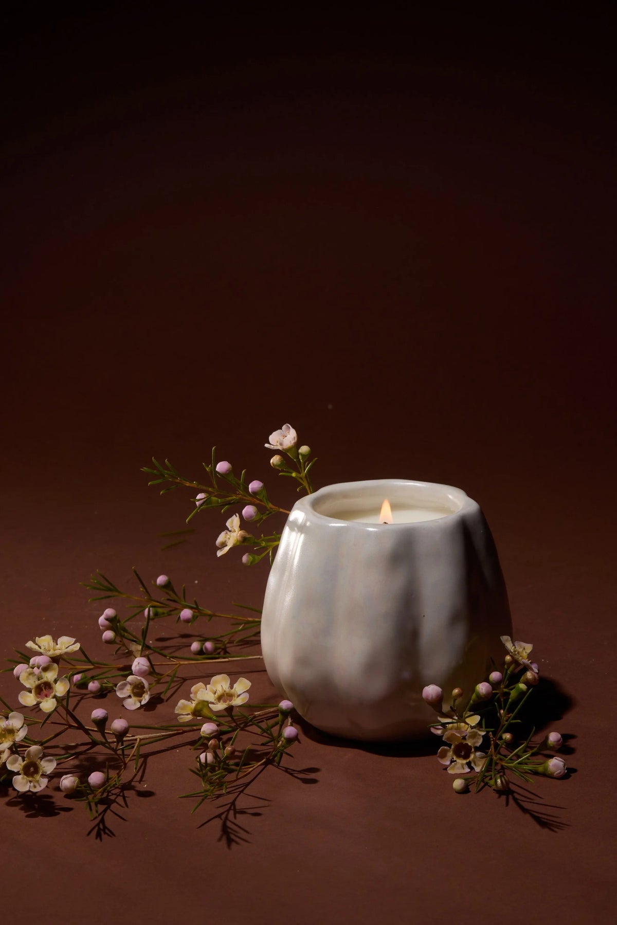 A lit white candle in a textured ceramic holder sits on a brown surface, surrounded by small pink and white flowers and green sprigs, against a dark brown background.