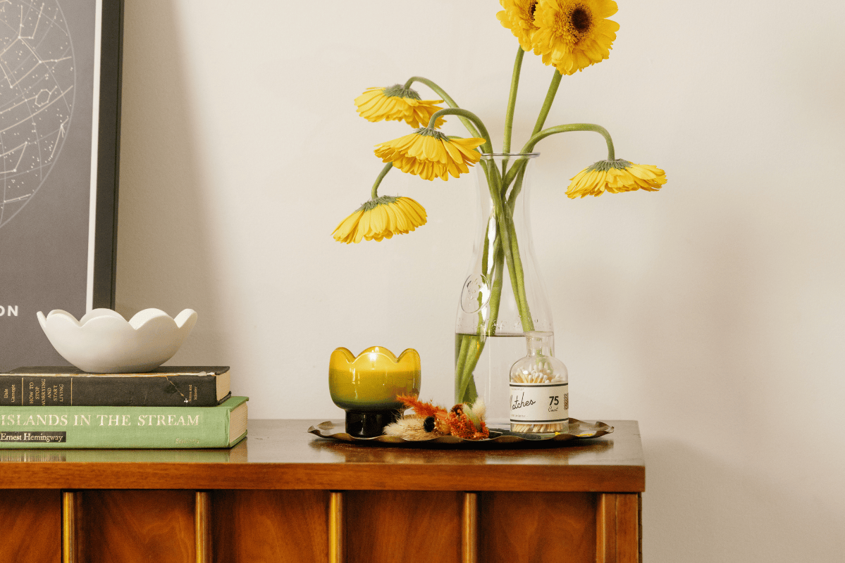 A wooden dresser holds stacked books, a white dish, a glass vase with yellow flowers, a yellow candle, a small bottle, and dried flowers on a tray, next to a framed artwork on the wall.