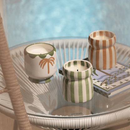 Three Bora Bora - Cabana 6.5 oz Candles and a book rest on a round glass-top wicker table by the pool, filling the air with inviting coastal scents.