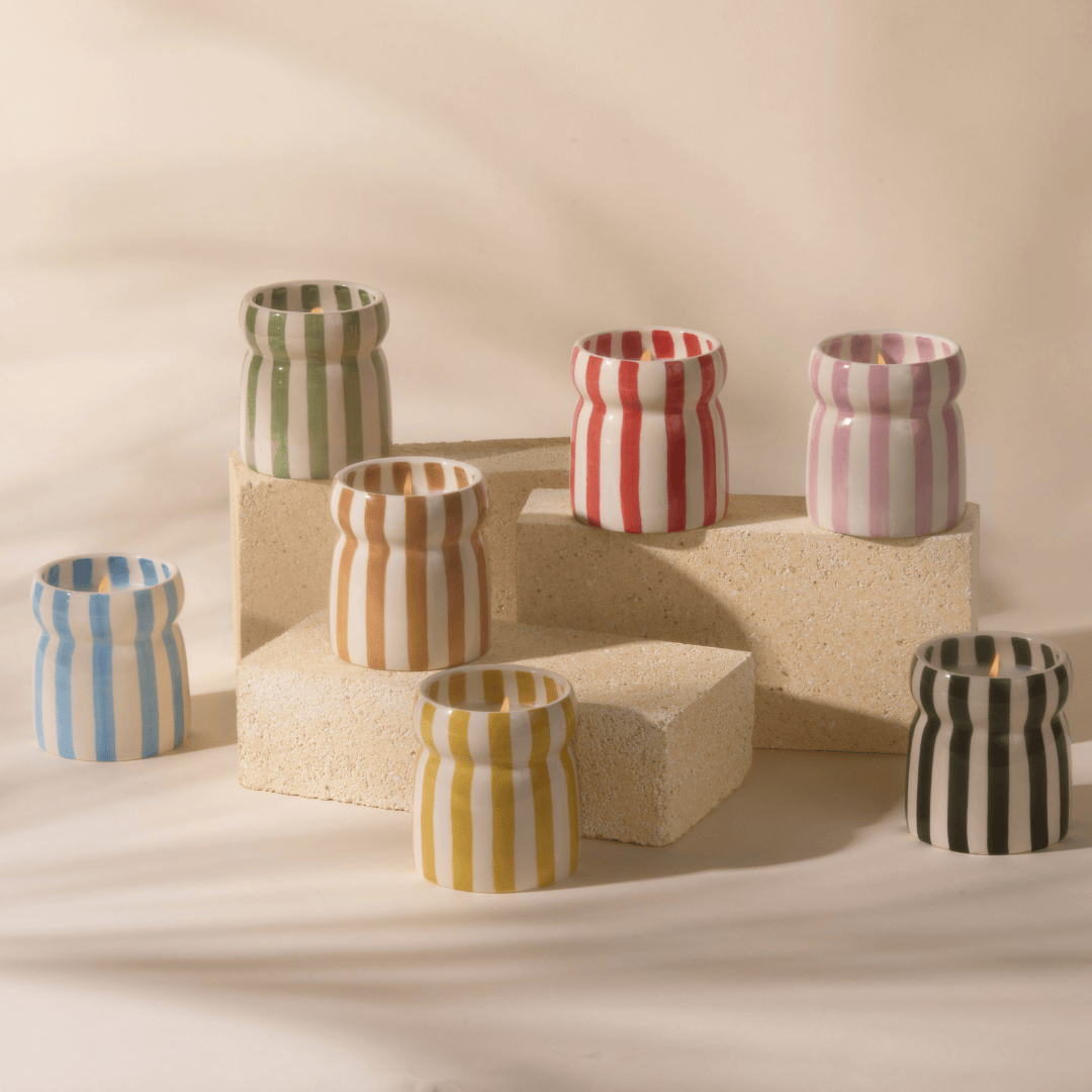 Seven Grecian Sand - Cabana 6.5 oz candles in striped ceramic vessels, each a different color, sit atop three cream rectangular blocks against a neutral backdrop, evoking a coastal-inspired collection.