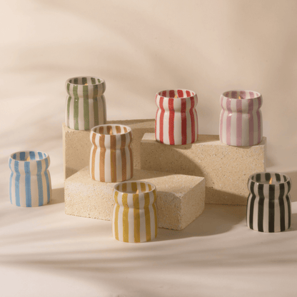 Seven Grecian Sand - Cabana 6.5 oz candles in striped ceramic vessels, each a different color, sit atop three cream rectangular blocks against a neutral backdrop, evoking a coastal-inspired collection.