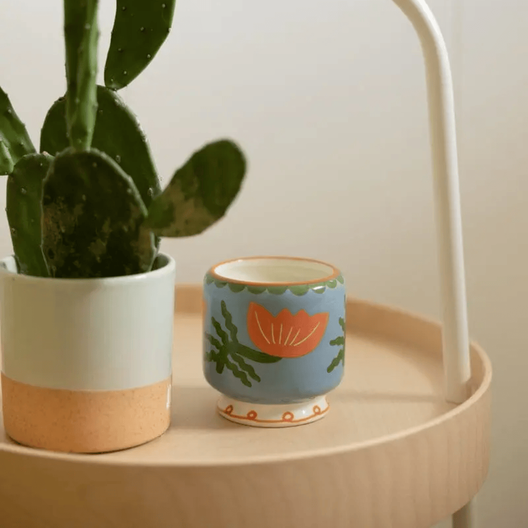 A green cactus in a white and tan pot sits next to a small blue mug with an orange flower design, paired with the Cactus Flower - A Dopo 8 oz Candle, all arranged on a round wooden surface.