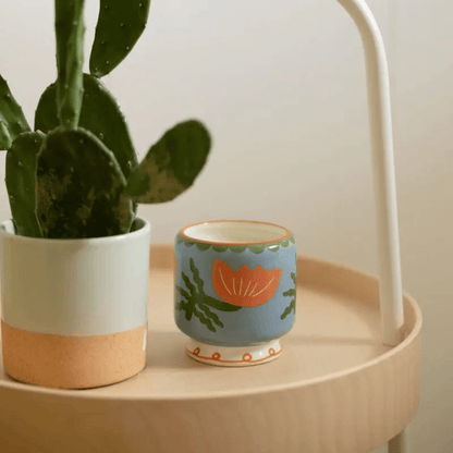 A green cactus in a white and tan pot sits next to a small blue mug with an orange flower design, paired with the Cactus Flower - A Dopo 8 oz Candle, all arranged on a round wooden surface.