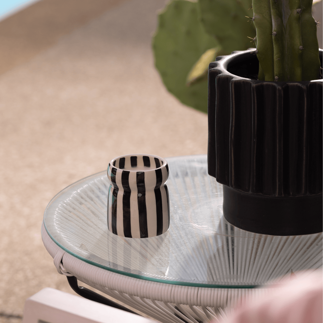 A Lava Stone - Cabana 6.5 oz Black Candle and a black planter with a cactus sit on a round glass table outdoors.