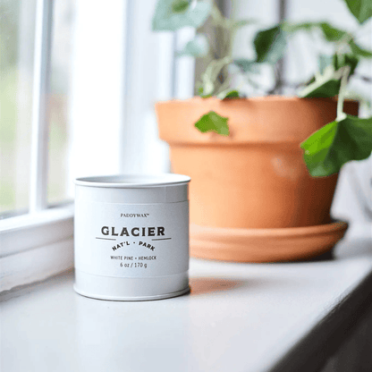 A Glacier - Parks 6 oz Tin Candle sits on a windowsill beside a green plant in a terracotta pot, filling your home with the fresh, National Park-inspired scent of this Parks Collection candle.