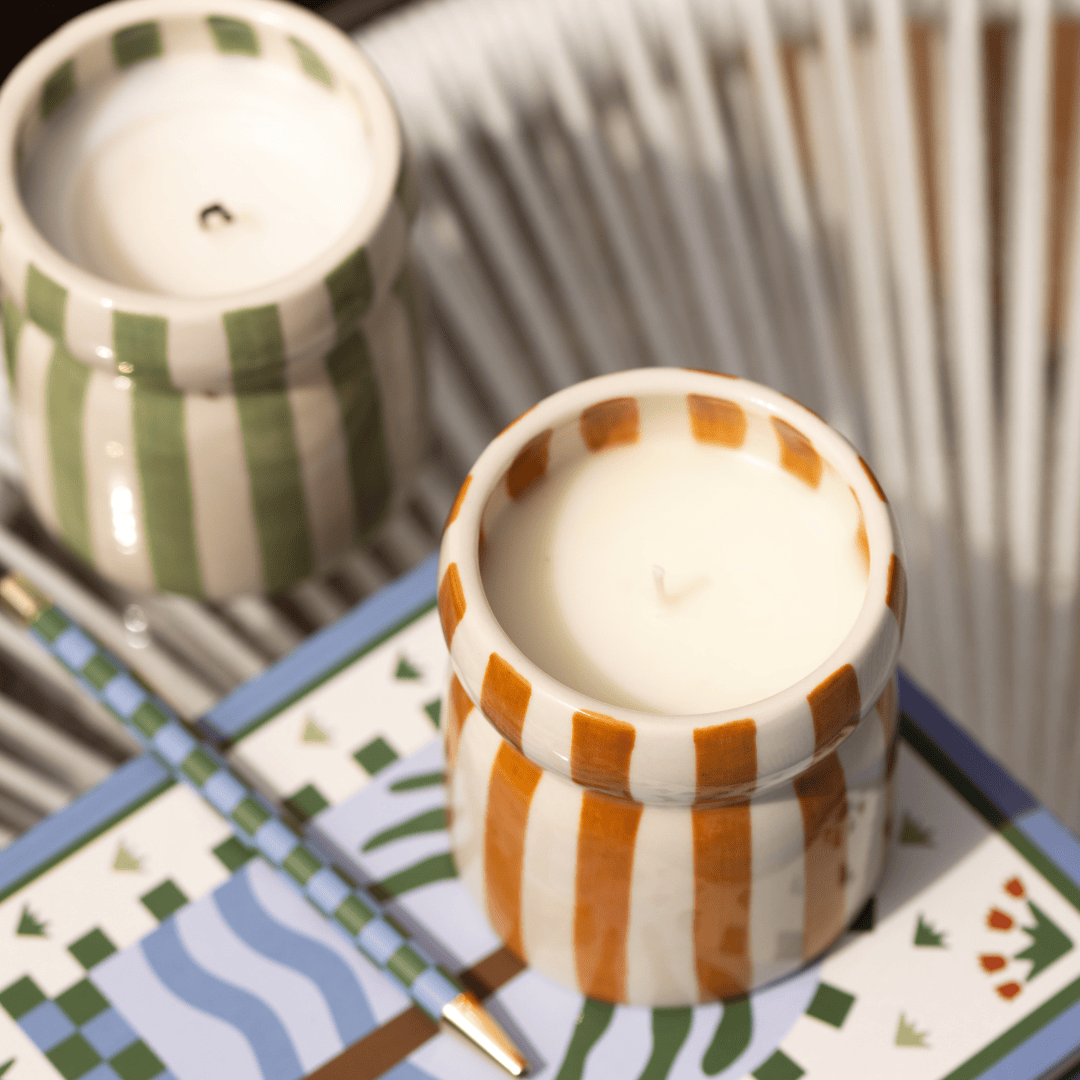 The Sienna Sunset - Cabana 6.5 oz candles, in hand-painted striped ceramic vessels—one orange, one green—add coastal scents while resting on a patterned book beside a striped pen atop a woven white surface.