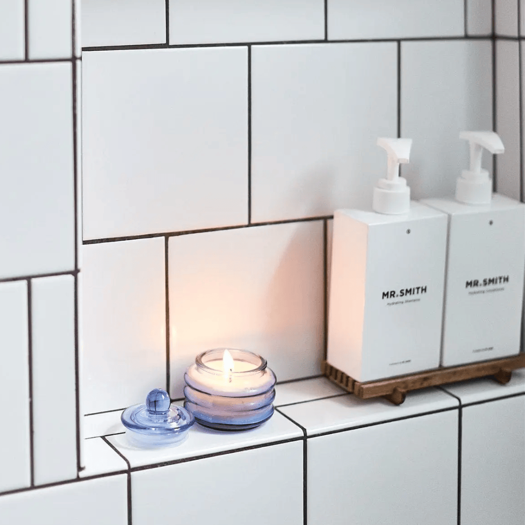 A Pepper + Plum - Beam 3 oz Candle in a blue glass holder casts ambient light beside two MR.SMITH white soap dispensers on a tiled bathroom shelf.