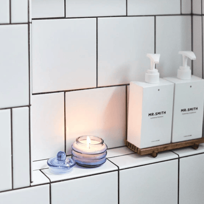 A Pepper + Plum - Beam 3 oz Candle in a blue glass holder casts ambient light beside two MR.SMITH white soap dispensers on a tiled bathroom shelf.