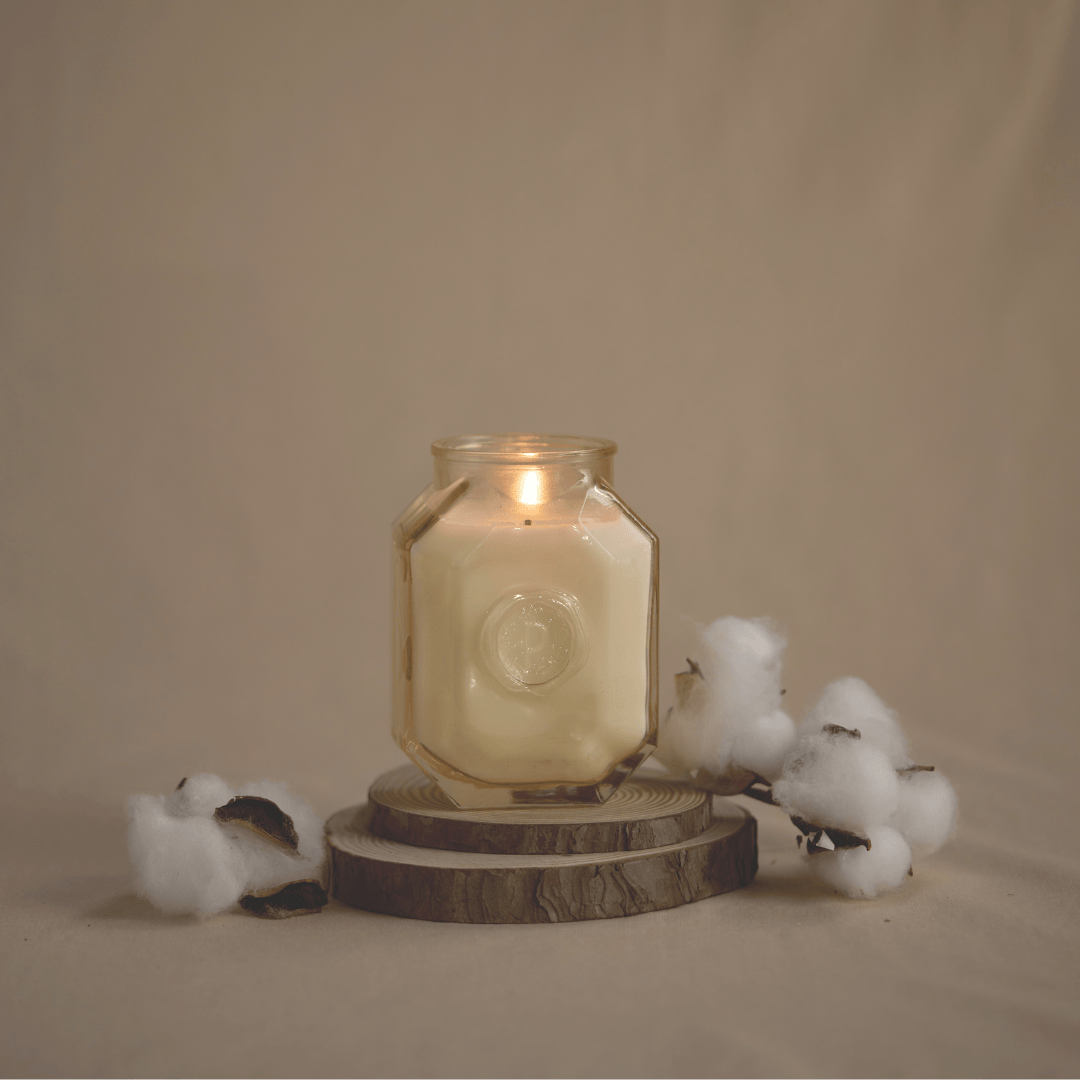 The Cotton + Teak - Botanica 8 oz Candle in a glass jar rests on wooden coasters, surrounded by cotton branches against a beige background, bringing an elegant touch to your home fragrance experience.