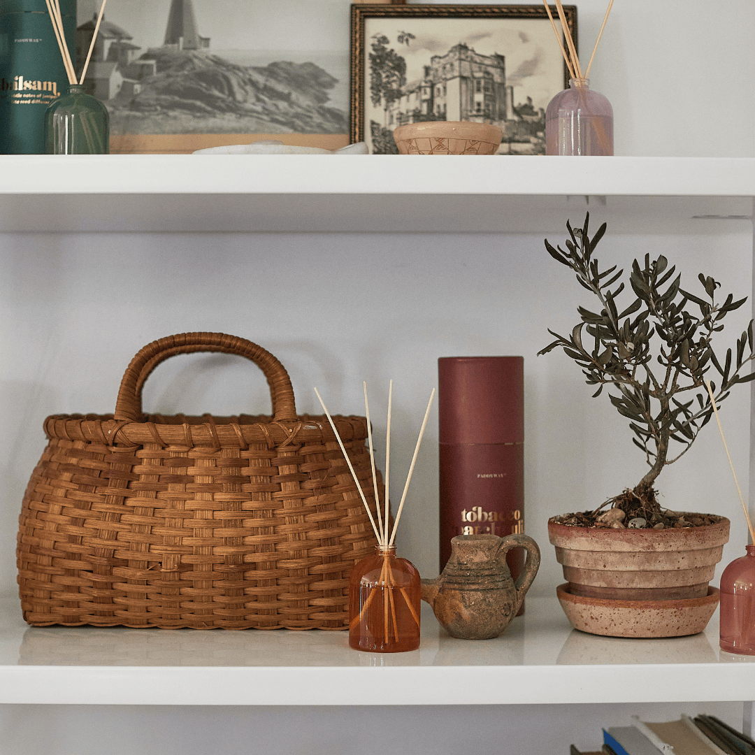 Two white shelves display a woven basket, potted plant, the Tobacco Patchouli 1.5 oz Diffuser with wooden reeds, framed photos, books, and pottery all arranged neatly.