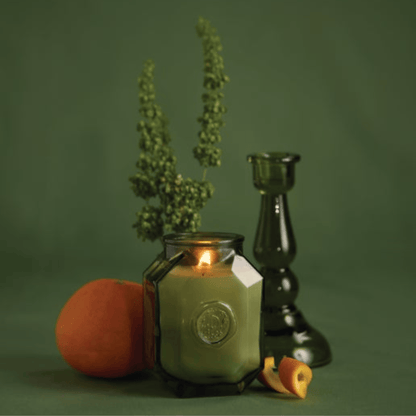 The Wild Neroli - Botanica 8 oz Candle glows in a glass holder beside a green candlestick, fresh orange, lush foliage, and orange petals—all arranged for an elegant home fragrance display against a green backdrop.