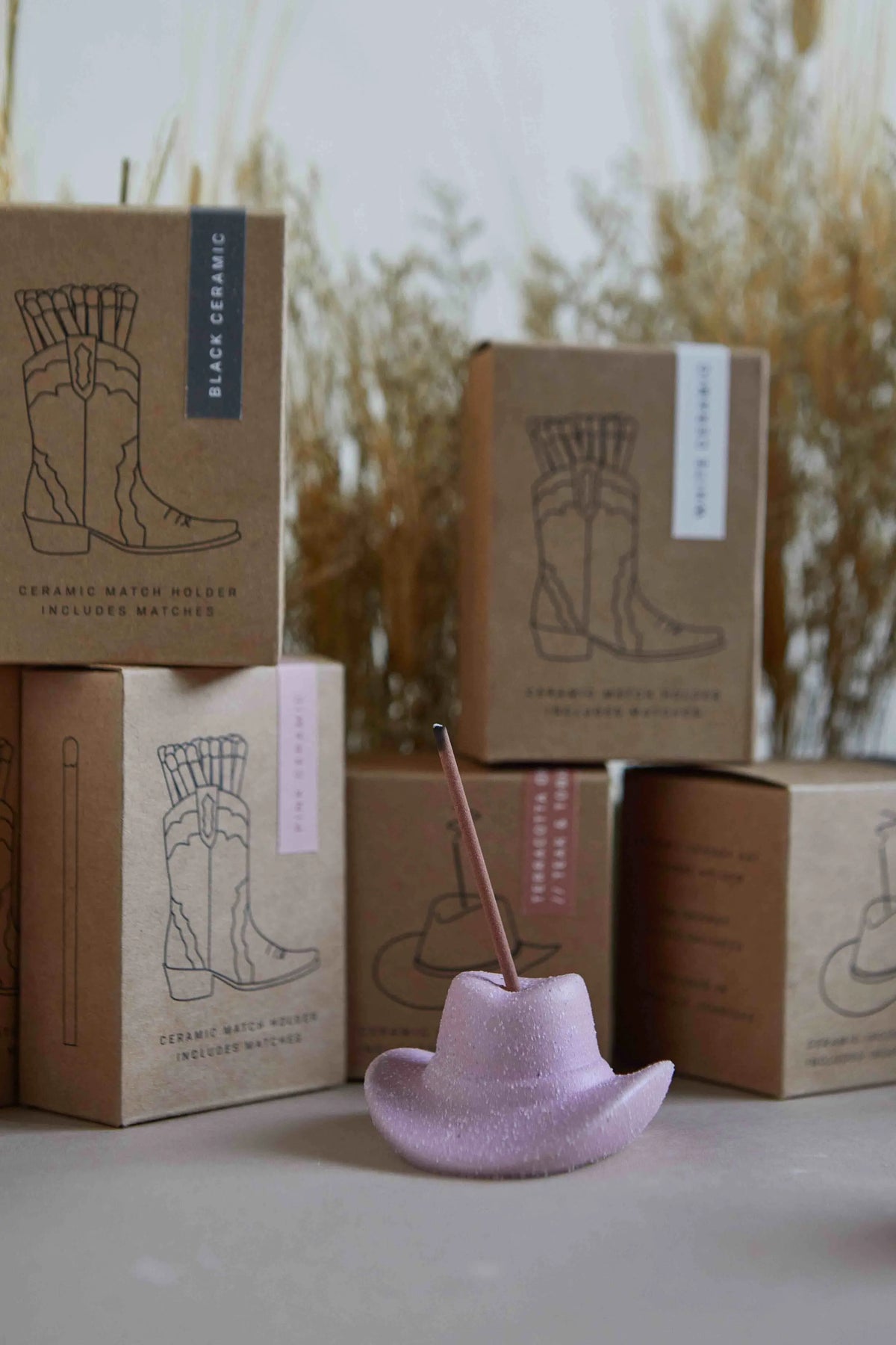 A pink cowboy hat-shaped holder, ideal for incense or matches, sits before brown boxes labeled Hinoki Wood - Incense Holder with boot graphics. Dried grass decorates the background, adding a quirky western touch.