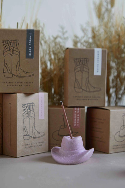 A pink cowboy hat-shaped holder, ideal for incense or matches, sits before brown boxes labeled Hinoki Wood - Incense Holder with boot graphics. Dried grass decorates the background, adding a quirky western touch.